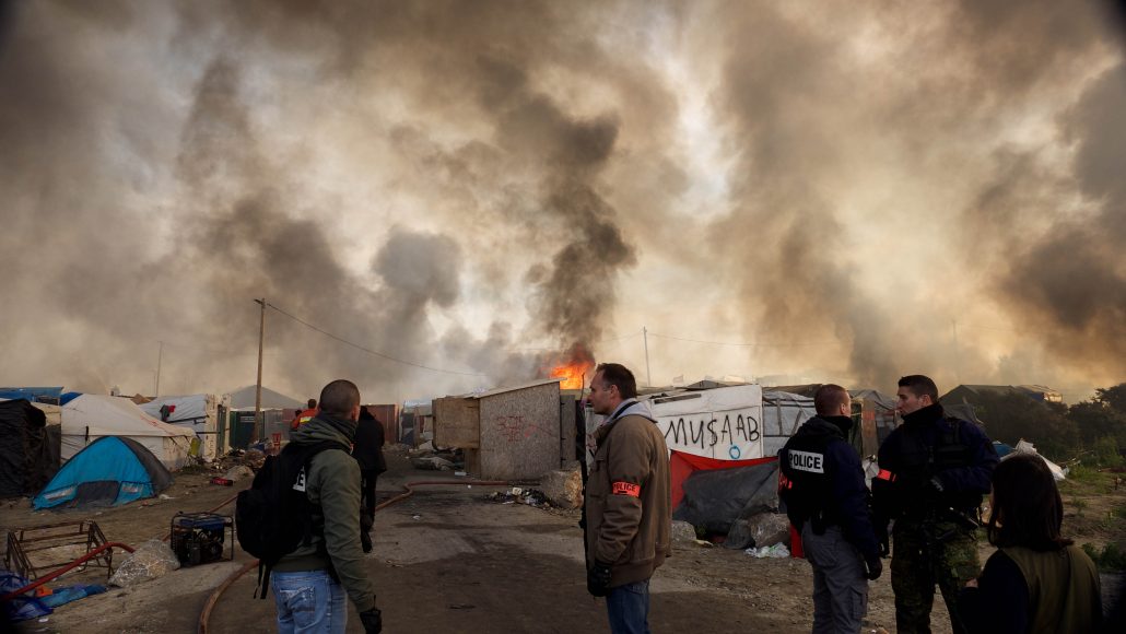 Police stand among burning shelters in the Calais refugee camp